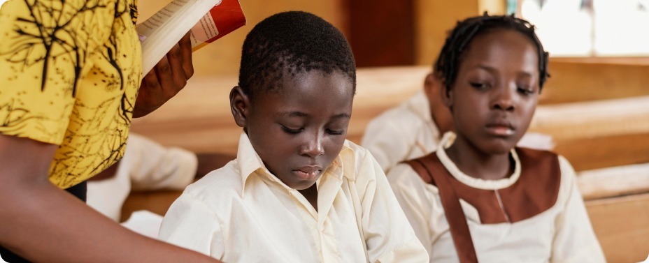 A supportive teacher mentoring a boy and a girl on a shared notebook, illustrating how school kits answer the UNESCO 2026 Education Monitoring Report during an April distribution drive.
