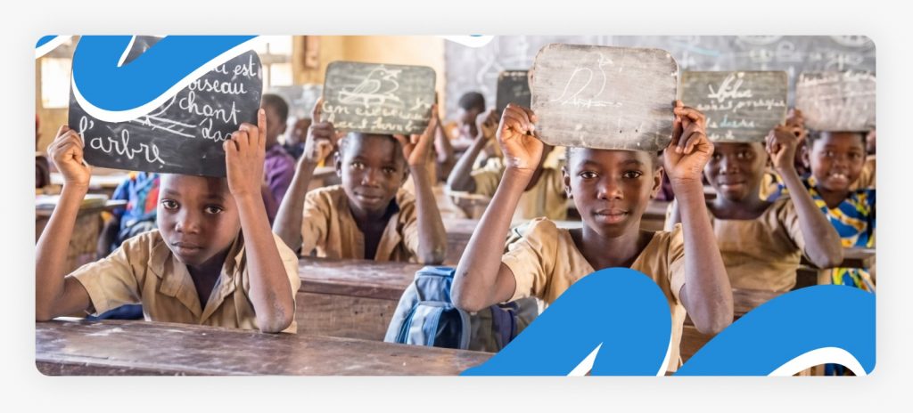Young students sitting in a classroom and proudly holding up individual small writing boards with chalk lessons written on them, showcasing the bright future unlocked by education for young orphans in Africa.